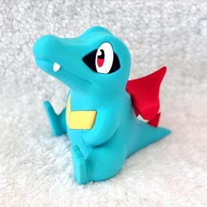 May include: A turquoise figurine of a cartoon crocodile with red wings and a yellow belly. The figurine has black eyes with white and red accents and is sitting down. The figurine is on a white, textured surface.
