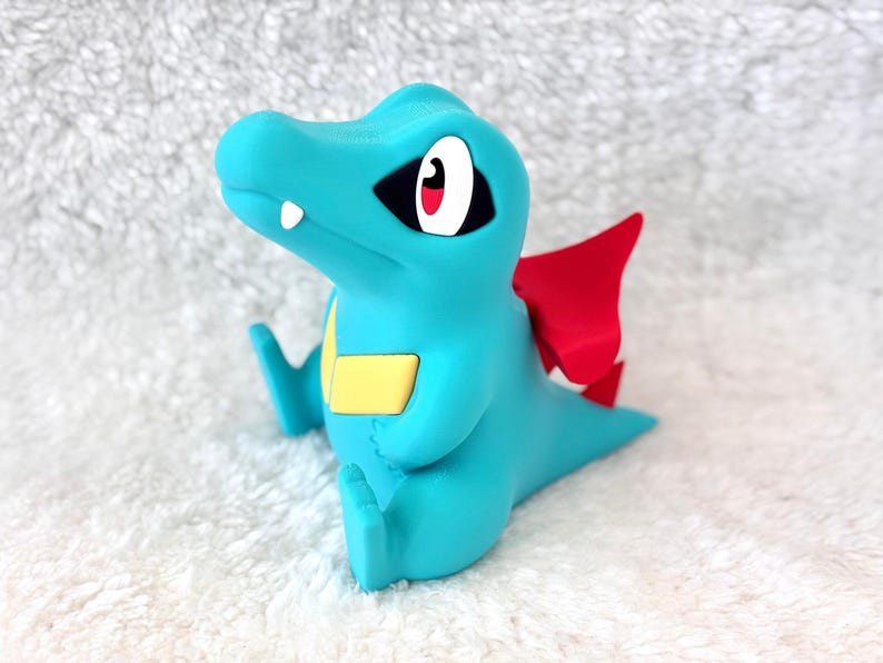 May include: A turquoise figurine of a cartoon crocodile with red wings and a yellow belly. The figurine has black eyes with white and red accents and is sitting down. The figurine is on a white, textured surface.