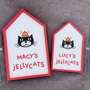 May include: Two house-shaped signs with red frames. The signs feature a black cat illustration wearing a hat, with the text "MACY'S JELLYCATS" and "LUCY'S JELLYCATS" in red. The larger sign is 26.2 cm tall, and the smaller one is 18.8 cm tall.