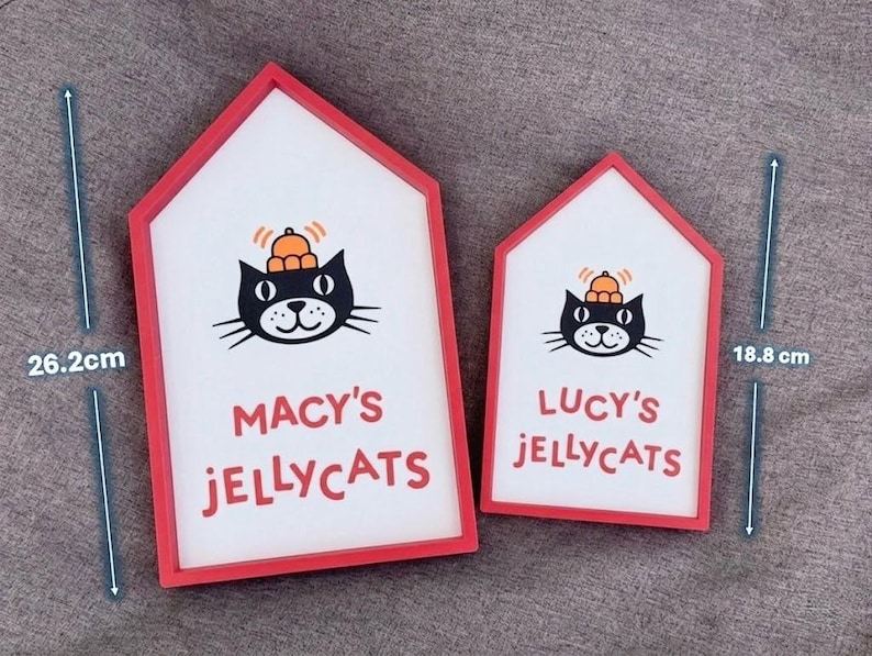 May include: Two house-shaped signs with red frames. The signs feature a black cat illustration wearing a hat, with the text "MACY'S JELLYCATS" and "LUCY'S JELLYCATS" in red. The larger sign is 26.2 cm tall, and the smaller one is 18.8 cm tall.