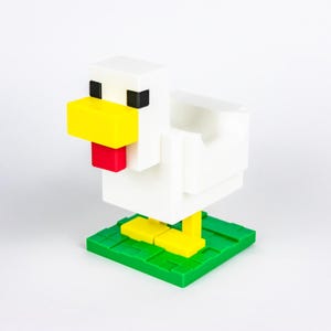 May include: A white, blocky chicken figurine with a yellow beak and feet, and a red wattle. The chicken stands on a green, square base. The eyes are black squares. The design is reminiscent of pixel art.