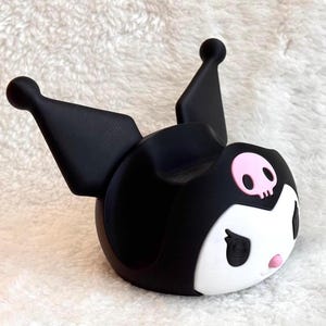 May include: A black and white character-themed phone stand featuring two tall pointed ears with rounded tips, a white face, black eyes, a small pink nose, and a pink skull emblem centered on the forehead. The stand has a hollow center.