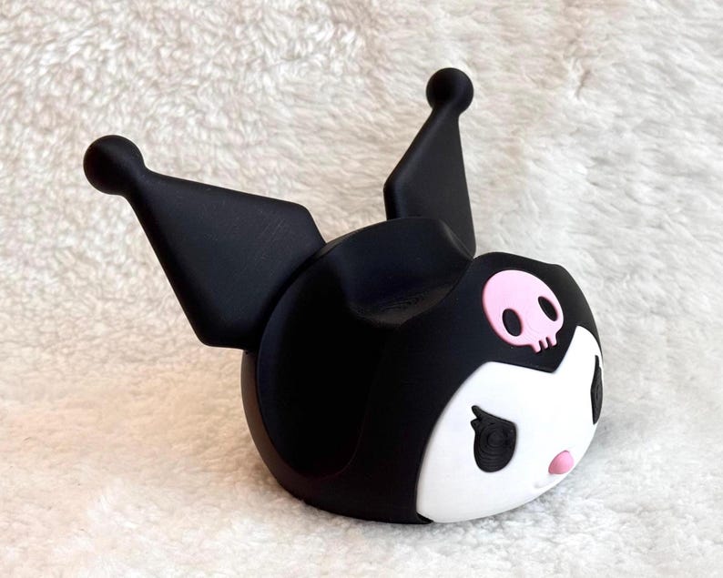 May include: A black and white character-themed phone stand featuring two tall pointed ears with rounded tips, a white face, black eyes, a small pink nose, and a pink skull emblem centered on the forehead. The stand has a hollow center.