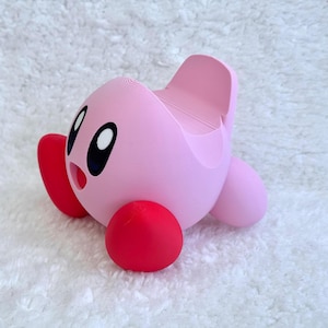 May include: A pink, spherical character figure with large black and white eyes and an open mouth. The figure has bright red, rounded feet and a smooth, matte finish. A recessed slot is integrated into the top back section of the character.