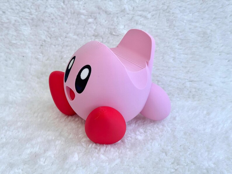 May include: A pink, spherical character figure with large black and white eyes and an open mouth. The figure has bright red, rounded feet and a smooth, matte finish. A recessed slot is integrated into the top back section of the character.