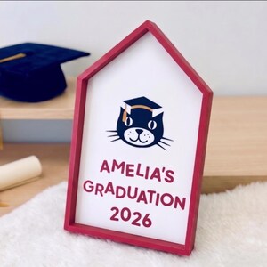 May include: A red-framed sign with the text "AMELIA'S GRADUATION 2026" and a cat wearing a graduation cap. A dark blue graduation cap with a gold tassel is visible in the background. The sign is on a white surface.