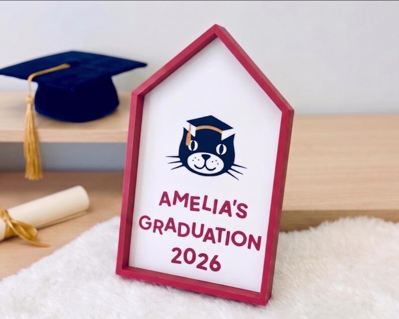 May include: A red-framed sign with the text "AMELIA'S GRADUATION 2026" and a cat wearing a graduation cap. A dark blue graduation cap with a gold tassel is visible in the background. The sign is on a white surface.