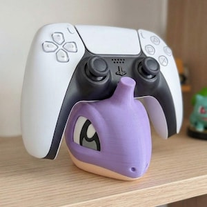 May include: A white and black PlayStation 5 controller rests above a purple and tan 3D-printed Pok&eacute;mon character. The controller is on a wooden shelf, with a green Pok&eacute;mon figure in the background.