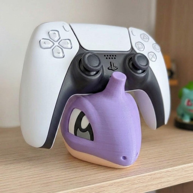 May include: A white and black PlayStation 5 controller rests above a purple and tan 3D-printed Pok&eacute;mon character. The controller is on a wooden shelf, with a green Pok&eacute;mon figure in the background.