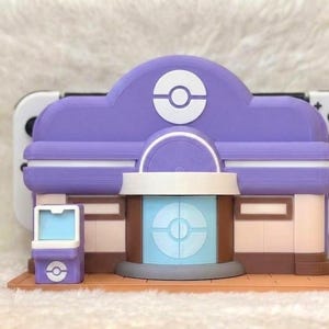 May include: A purple and white 3D-printed model of a building with a circular logo on the roof and front. The model has a light blue and brown facade. A white game console is behind the model.