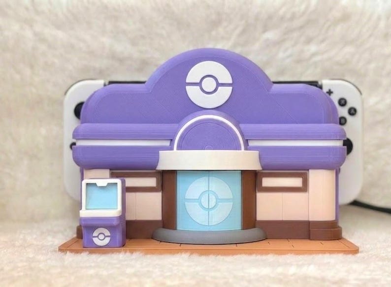 May include: A purple and white 3D-printed model of a building with a circular logo on the roof and front. The model has a light blue and brown facade. A white game console is behind the model.