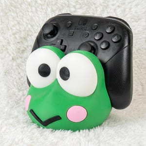 Puede incluir: Un soporte para mando con forma de rana verde que presenta grandes ojos blancos con pupilas negras, mejillas circulares rosas y una boca negra en forma de V. Un mando negro de Nintendo Switch Pro descansa en el soporte, mostrando los botones.