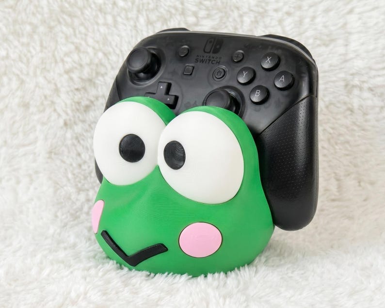 Puede incluir: Un soporte para mando con forma de rana verde que presenta grandes ojos blancos con pupilas negras, mejillas circulares rosas y una boca negra en forma de V. Un mando negro de Nintendo Switch Pro descansa en el soporte, mostrando los botones.