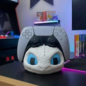 May include: A white and blue dragon-themed PlayStation 5 controller stand. The stand features a dragon head with blue eyes and a grey controller on top. The stand is on a black surface.