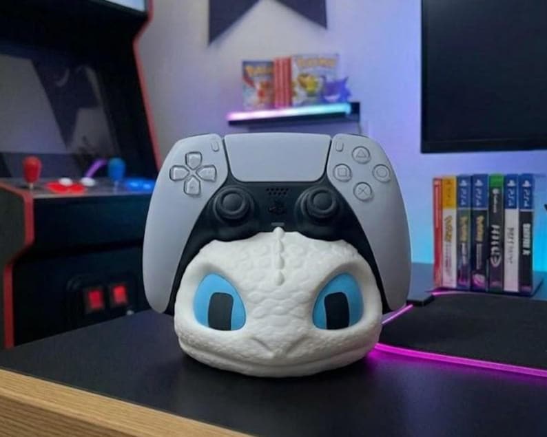 May include: A white and blue dragon-themed PlayStation 5 controller stand. The stand features a dragon head with blue eyes and a grey controller on top. The stand is on a black surface.