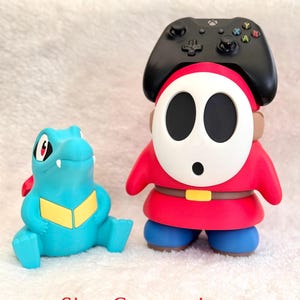 May include: A blue and yellow Totodile figurine next to a red, white, and blue Shy Guy figurine wearing a black Xbox controller on its head. The text "Size Comparison" is printed in red at the bottom of the image, showing the scale of the items.