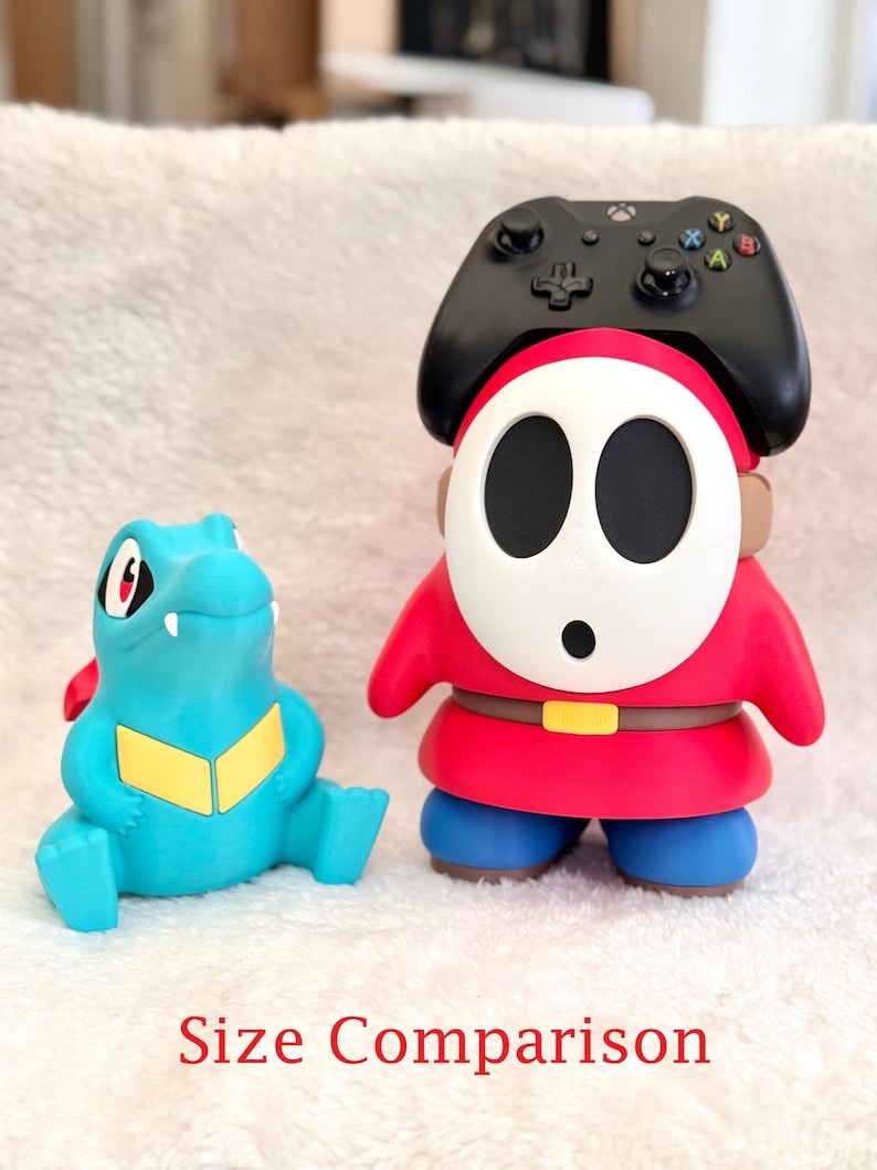 May include: A blue and yellow Totodile figurine next to a red, white, and blue Shy Guy figurine wearing a black Xbox controller on its head. The text "Size Comparison" is printed in red at the bottom of the image, showing the scale of the items.