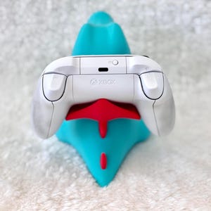 May include: A white Xbox controller rests on a teal and red stand. The controller has a sleek design with the Xbox logo visible. The stand is shaped like a stylized creature, providing a vibrant contrast to the controller.