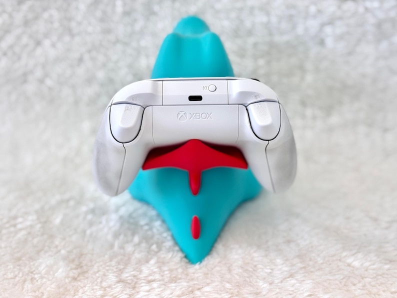 May include: A white Xbox controller rests on a teal and red stand. The controller has a sleek design with the Xbox logo visible. The stand is shaped like a stylized creature, providing a vibrant contrast to the controller.