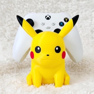 May include: A yellow Pikachu character figure serving as a controller stand, positioned in front of a white wireless gaming controller with black analog sticks and a central Xbox logo button, featuring red cheek circles and black-tipped ears.