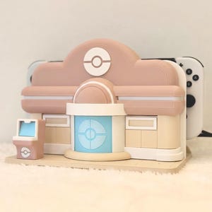 May include: A pastel pink and beige gaming console dock shaped like a building with a central blue door, a white circular emblem, and a small matching arcade cabinet accessory. The dock holds a white gaming console with black buttons visible on the side.