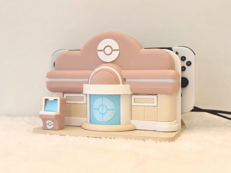 May include: A pastel pink and beige gaming console dock shaped like a building with a central blue door, a white circular emblem, and a small matching arcade cabinet accessory. The dock holds a white gaming console with black buttons visible on the side.