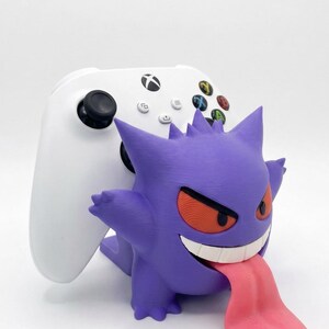 May include: A white wireless game controller resting on a purple Gengar character stand. The stand features the character with spiky ears, orange eyes, a wide white grin, and a long pink tongue extending outward from the mouth area.