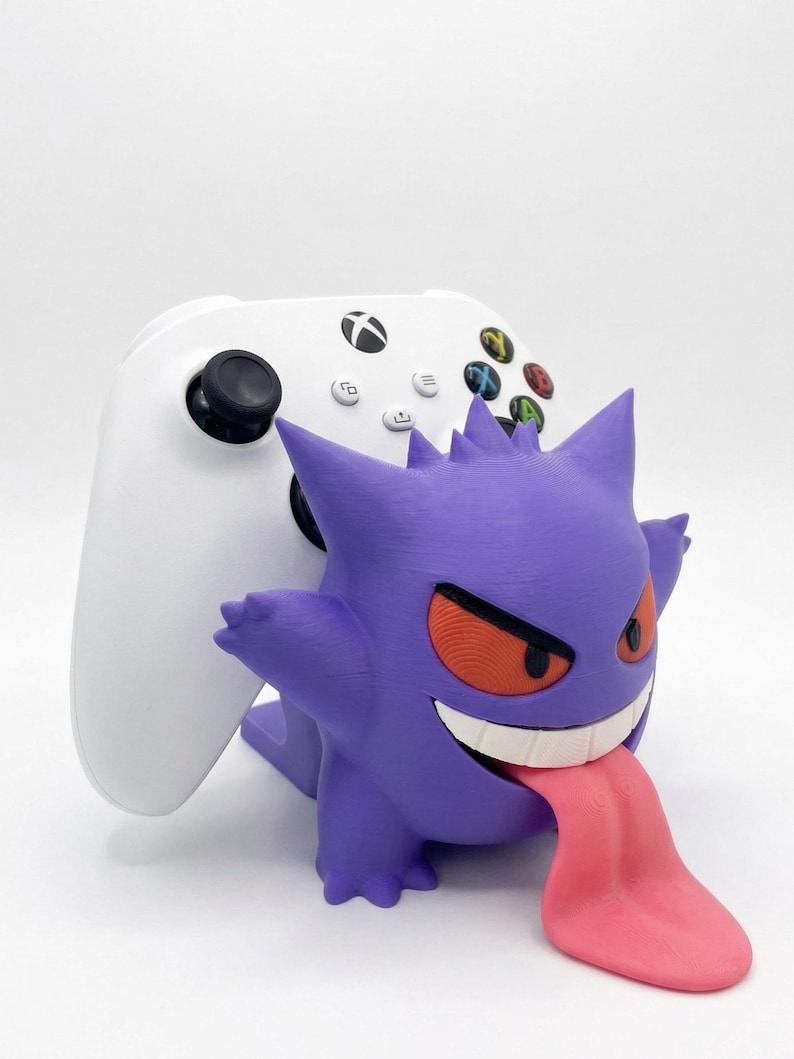 May include: A white wireless game controller resting on a purple Gengar character stand. The stand features the character with spiky ears, orange eyes, a wide white grin, and a long pink tongue extending outward from the mouth area.