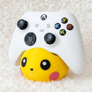 Peut inclure: Une manette de jeu sans fil blanche posée sur un support jaune en forme de personnage. Le support présente des yeux noirs, un petit nez et des joues roses circulaires. La manette possède des joysticks noirs, une croix directionnelle et des boutons.
