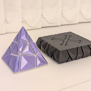 May include: A geometric puzzle in the shape of a pyramid, in shades of lavender, sits next to a black, angular device with a cross-shaped design. The pyramid is composed of triangular and quadrilateral facets. The device has a power cord.