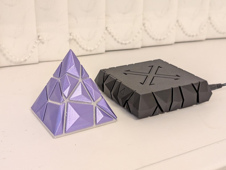 May include: A geometric puzzle in the shape of a pyramid, in shades of lavender, sits next to a black, angular device with a cross-shaped design. The pyramid is composed of triangular and quadrilateral facets. The device has a power cord.