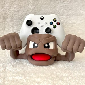 May include: A white Xbox controller with black buttons and colorful action buttons is partially visible behind a brown cartoon figure with a red mouth and clenched fists. The figure appears to be a Geodude from the Pokemon series.