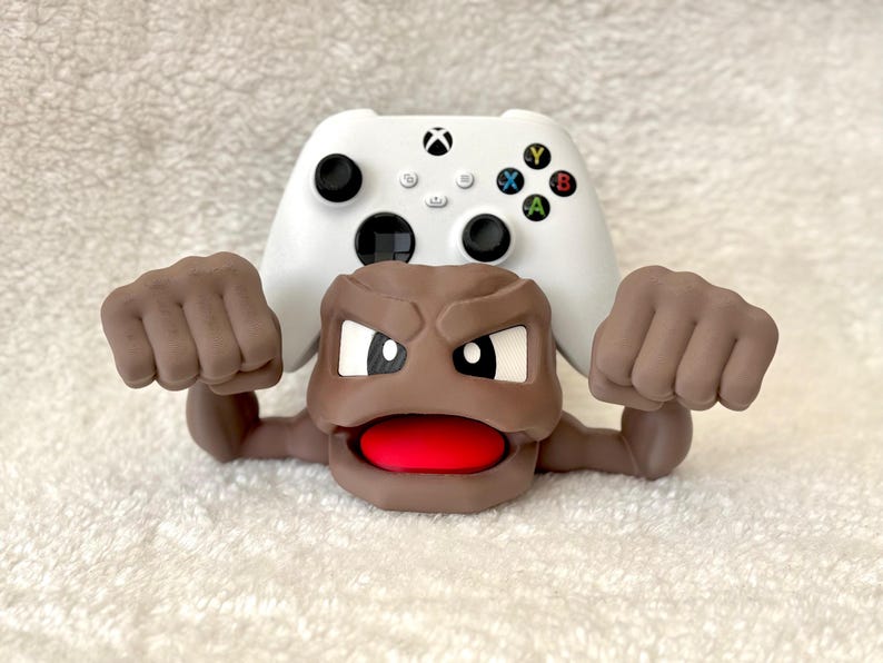 May include: A white Xbox controller with black buttons and colorful action buttons is partially visible behind a brown cartoon figure with a red mouth and clenched fists. The figure appears to be a Geodude from the Pokemon series.