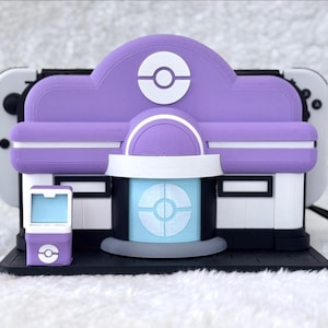 May include: A purple and white 3D-printed model of a Pok&eacute;mon Center, with a matching small device. The model features a Pok&eacute; Ball design on the roof and a light blue circular design on the front. The model is designed to hold a Nintendo Switch.