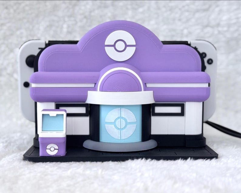 May include: A purple and white 3D-printed model of a Pok&eacute;mon Center, with a matching small device. The model features a Pok&eacute; Ball design on the roof and a light blue circular design on the front. The model is designed to hold a Nintendo Switch.
