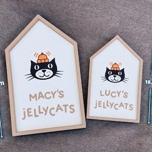 May include: Two house-shaped wooden signs with a white background and a cartoon cat illustration. The larger sign is 26.2 cm tall and reads "MACY'S JELLYCATS". The smaller sign is 18.8 cm tall and reads "LUCY'S JELLYCATS".