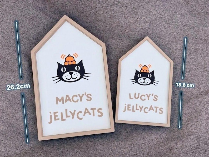 May include: Two house-shaped wooden signs with a white background and a cartoon cat illustration. The larger sign is 26.2 cm tall and reads "MACY'S JELLYCATS". The smaller sign is 18.8 cm tall and reads "LUCY'S JELLYCATS".