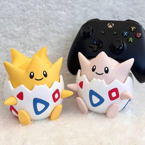 May include: Two round character figures, one yellow and one pale pink, each emerging from a white eggshell base with blue and red triangle patterns. A black wireless game controller rests on top of the pale pink figure's head.