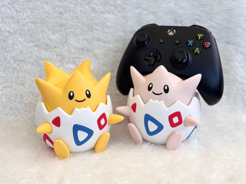 May include: Two round character figures, one yellow and one pale pink, each emerging from a white eggshell base with blue and red triangle patterns. A black wireless game controller rests on top of the pale pink figure's head.