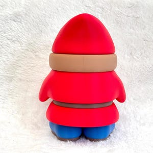 May include: A gnome figurine featuring a bright red pointed hat, a tan band around the hat, a red tunic with a brown belt, and blue boots. The figure has a smooth, matte finish and is shown from the back, displaying a solid, uniform color scheme.