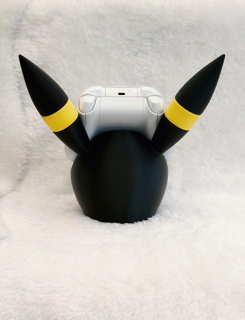 May include: A black controller stand featuring two tall, pointed ears with yellow bands, designed to hold a white game controller. The stand has a smooth, rounded base and the ears extend upward, mimicking the appearance of a creature's head.