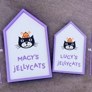 May include: Two house-shaped signs with a white background and a purple frame. Each sign features a black cat illustration with an orange hat. The larger sign reads "MACY'S JELLYCATS" and is 26.2 cm tall. The smaller sign reads "LUCY'S JELLYCATS" and is 18.8 cm tall.
