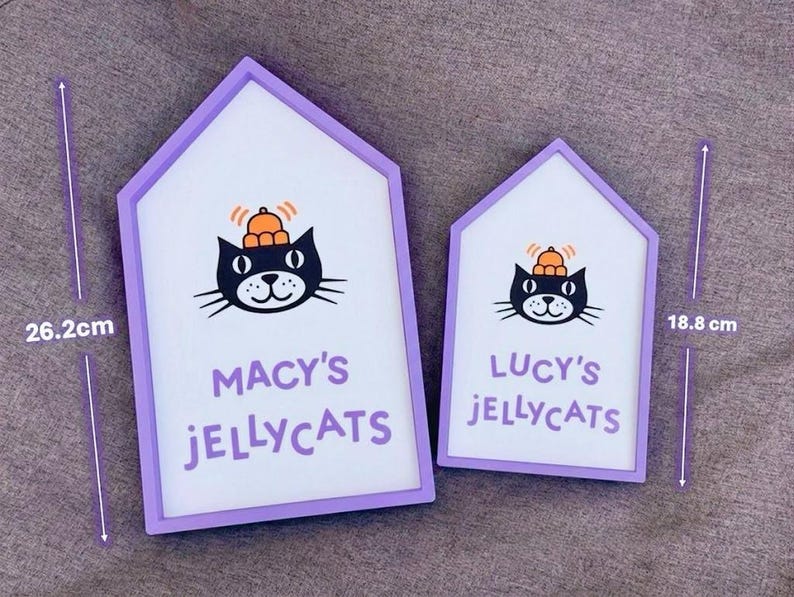 May include: Two house-shaped signs with a white background and a purple frame. Each sign features a black cat illustration with an orange hat. The larger sign reads "MACY'S JELLYCATS" and is 26.2 cm tall. The smaller sign reads "LUCY'S JELLYCATS" and is 18.8 cm tall.