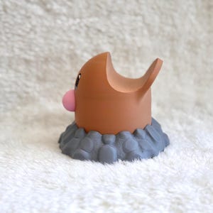 May include: A brown, rounded figurine with a small pink nose and a large, open-mouthed expression, emerging from a textured, gray, rock-like base. The character has a smooth, matte finish and a distinct, curved shape that resembles a burrowing creature.
