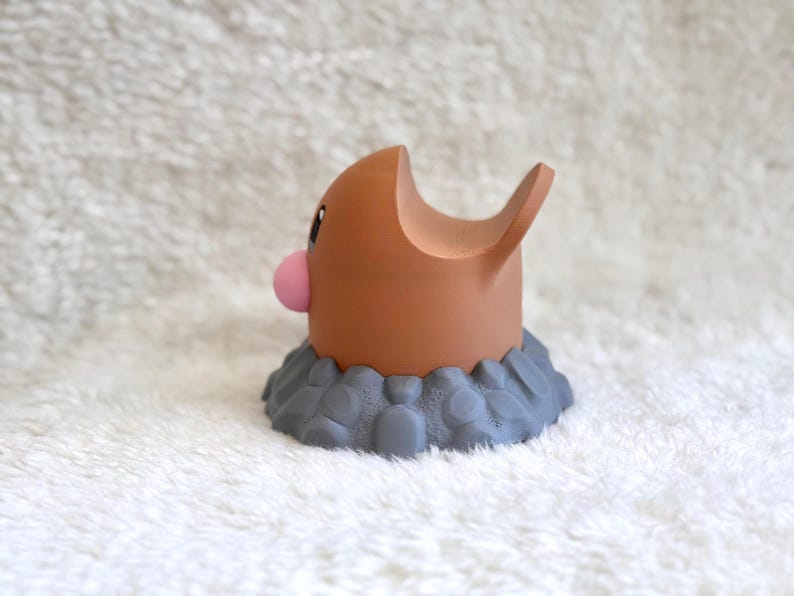 May include: A brown, rounded figurine with a small pink nose and a large, open-mouthed expression, emerging from a textured, gray, rock-like base. The character has a smooth, matte finish and a distinct, curved shape that resembles a burrowing creature.