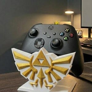May include: A white and gold 3D-printed stand featuring the iconic Triforce symbol from The Legend of Zelda, with a black Xbox controller in the background. The stand is designed to hold the controller, showcasing a gaming accessory.