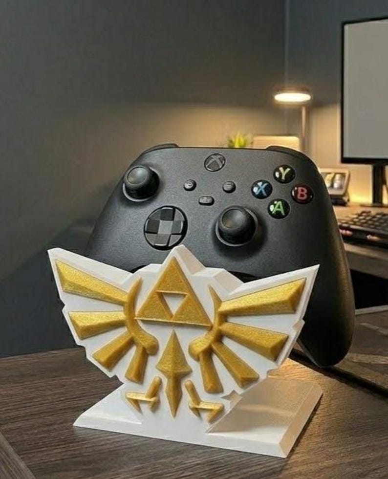May include: A white and gold 3D-printed stand featuring the iconic Triforce symbol from The Legend of Zelda, with a black Xbox controller in the background. The stand is designed to hold the controller, showcasing a gaming accessory.