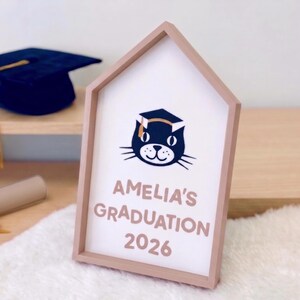May include: A wooden framed sign with a house-shaped design. The sign features the text "AMELIA'S GRADUATION 2026" and a cat wearing a graduation cap. A graduation cap and diploma are visible in the background.