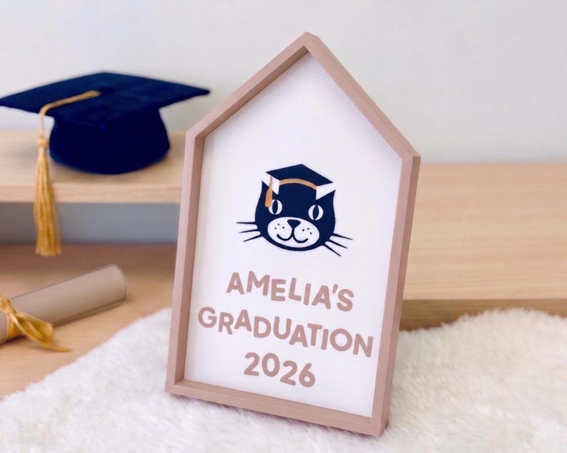 May include: A wooden framed sign with a house-shaped design. The sign features the text "AMELIA'S GRADUATION 2026" and a cat wearing a graduation cap. A graduation cap and diploma are visible in the background.