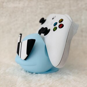 Puede incluir: Un soporte de mando azul claro con un mando Xbox blanco. El soporte tiene una forma redondeada con detalles en blanco y negro. El mando es blanco con botones de colores.
